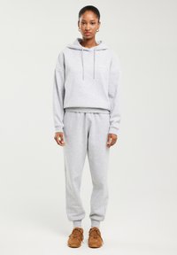 Kobieta stoi zwrócona przodem, ubrana w jasnoszarą bluzę z kapturem i spodnie dresowe, na nogach brązowe trampki, na jednolitym jasnym tle.