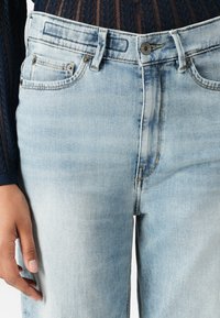 Jeans in denim azzurro chiaro con vita alta, design a cinque tasche, cuciture a contrasto e leggera scoloritura. Chiusura con bottone e zip.