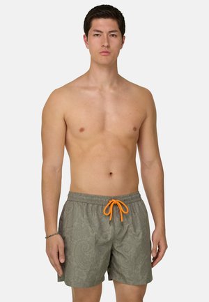 Joven de pie de frente, sin camiseta, con pantalones cortos de baño grises con estampado paisley y cordón naranja brillante.