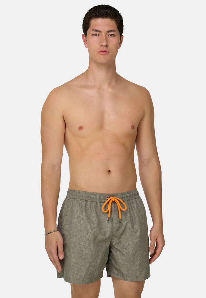 Jeune homme debout face à nous, torse nu, portant un short de bain gris à motif paisley avec un cordon orange vif.