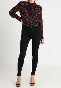 Blusa negra con estampado de flores rojas, cuello con volantes, y mangas largas, combinada con unos jeans negros ajustados y tacones negros con tiras.