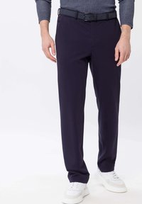 Homme portant un pantalon bleu marine foncé, une ceinture noire, des baskets blanches et une chemise à manches longues à motifs, se tenant sur un fond blanc.