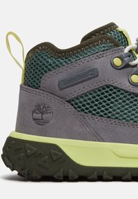 Stivale da trekking in suede grigio con tessuto in rete, dettagli verdi e gialli. Presenta una suola in gomma nera texturizzata e il logo Timberland.