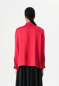 Blouse rouge légère avec une coupe décontractée, des manches longues et un col. Le dos présente un ourlet droit et des fentes latérales discrètes.