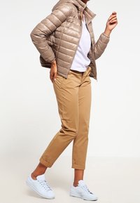 Brun pufferjacka, lätt med horisontell söm; ljusgrå topp; beige uppvikta byxor; vita sneakers med blå accenter.