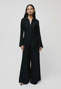 Conjunto texturizado em preto, composto por uma camisa de manga longa com botão e calças de perna ampla, apresentando riscas verticais subtis e um design estruturado.