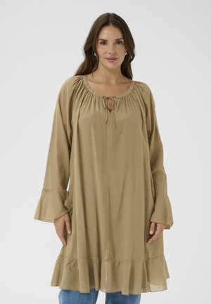 Kvinde med langt brunt hår iført en løs tan-farvet langærmet kjole med flæsekant og snor ved halsudskæringen, stående foran en ensfarvet baggrund.