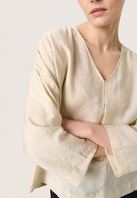 Blusa beige leggera con scollatura a V e maniche larghe. Il materiale sembra morbido, con una texture leggermente stropicciata e un design minimalista.