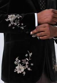 Black velvet jacket featuring intricate floral embroidery in cream, beige, and brown on the sleeves and front, with a smooth texture.
