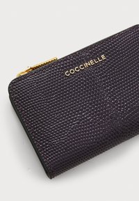 Tume lilla rahakott, millel on reljeefne tekstuur, kuldse tooni riistvara ja "Coccinelle" brändi logo. Lukk on paigaldatud ülemisele servale.