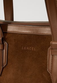 Sac à main en cuir suédé marron avec des accents en cuir lisse, une fermeture éclair et un logo embossé "Lancel Paris" à l'avant. Bandoulières robustes incluses.