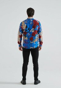 Chemise à manches longues bleue avec des motifs floraux et animaliers, mettant en avant des roses rouges et des taches de léopard, associée à un pantalon slim noir.