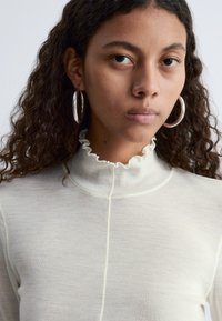 Helle beige Langarmbluse mit einem rüschierten Stehkragen, gerippter Textur und einer zentralen Naht. Silberne Ohrhänger ergänzen den Look.