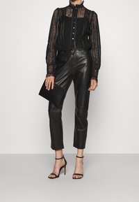 Blouse en dentelle noire à manches longues et col montant, associée à un pantalon en cuir noir ajusté et des sandales à talons noires. Pochette noire à la main.