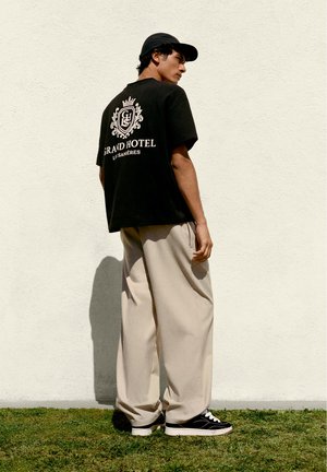 Jeune homme portant un T-shirt noir « Grand Hotel Les Sanières », un pantalon large beige, une casquette noire et des baskets noires, debout contre un mur blanc.