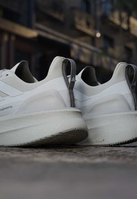 Coppia di scarpe da ginnastica bianche con suole spesse e linguette grigie posate su una superficie di pietra all'aperto, alla luce tenue.