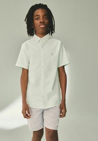 Világoszöld, rövid ujjú, gombos ing, gallérral és kis logóval, világosszürke nadrággal párosítva; sima anyag és laza fazon.