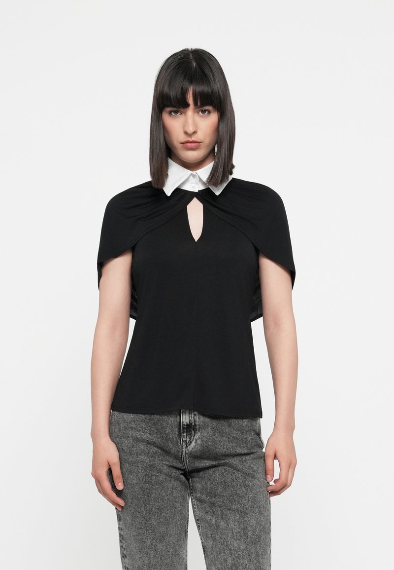 Schwarzes Oberteil mit kurzen, drapierten Ärmeln, einem weißen Kragen und einem Schlüssel-Ausschnitt. Getragen mit grauen, hochtaillierten Jeans. Glatte Textur insgesamt.