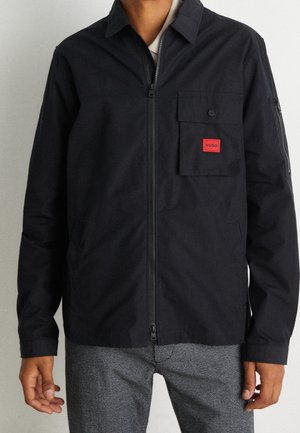 Homme portant une veste noire zippée avec une poche poitrine munie d'une étiquette rouge, associée à un pantalon gris à carreaux, debout devant un fond uni.