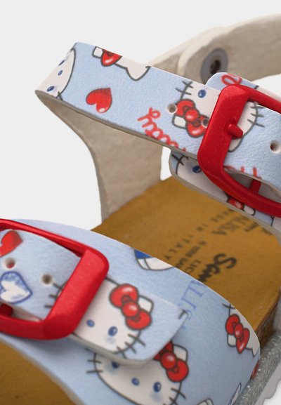 Πέδιλο παιδικό με λευκές λουρίδες που έχουν πρόσωπα Hello Kitty και κόκκινη καρδιά, ασφαλισμένο με κόκκινες πλαστικές αγκράφες.