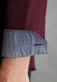 Burgunderfarbener Hemdmanschette mit einem gemusterten blauen Innenfutter, das kleine weiße Kreise und Blumen zeigt. Auffällige Stickerei-Akzentierung near der Kante.