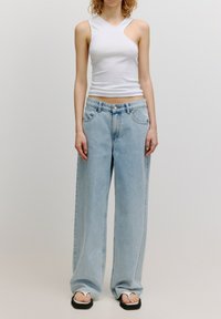 Weißes ärmelloses Oberteil mit asymmetrischem Ausschnitt, kombiniert mit hellblauen weiten Jeans. Die Fußbekleidung besteht aus schwarz-weißen Flip-Flops.