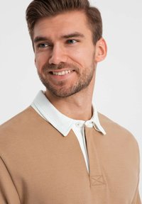 Beige Langarmoberteil mit einem weißen Hemdkragen, versehen mit einer Knopfleiste mit einer gestickten Akzentuierung. Glatte Stofftextur.