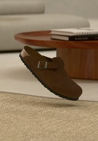 Καφέ σουέντ παπούτσι slip-on με λουράκι και αγκράφα που αιωρείται πάνω από μπεζ χαλί σε μοντέρνο σαλόνι με ξύλινο τραπεζάκι καφέ και βιβλία.