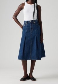 Falda de mezclilla en azul oscuro, hasta la rodilla, con pliegues y cintura alta, combinada con una camiseta sin mangas blanca y bailarina negra. Textura suave.