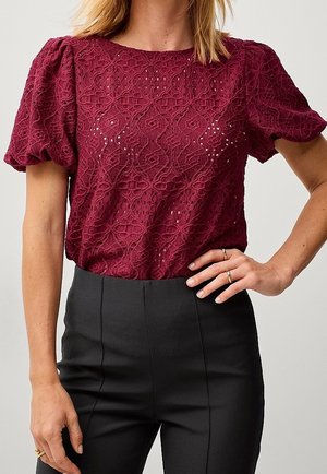 Kobieta nosząca bordową koronkową bluzkę z krótkimi bufkami i czarne spodnie z wysokim stanem, ręka oparta na biodrze z pierścionkami.