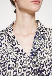 Blusa com padrão de leopardo, apresentando um decote com gola, tecido leve e uma mistura de cores bege, verde oliva e azul marinho com uma textura suave.