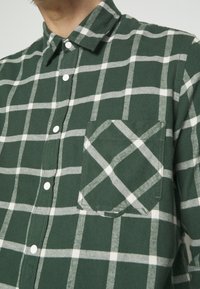 Chemise à carreaux vert et blanc à boutons, avec une poche poitrine avant et un col, portée par une personne visible du cou jusqu'au milieu du torse.