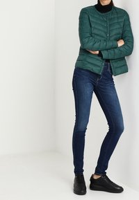 Veste coupe-vent verte avec un design matelassé, associée à un jean skinny noir et des baskets noires. L'ensemble présente une silhouette ajustée.
