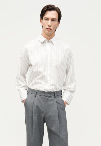 Άνδρας με λευκό πουκάμισο με ριγέ κουμπιά, φορεμένο μέσα σε γκρι πλισέ παντελόνι, με τα χέρια στις τσέπες, κοιτάζοντας πλάγια σε μονόχρωμο φόντο.