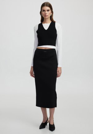 Top corto negro con relieve, superpuesto sobre una camisa blanca de manga larga, combinado con una falda midi negra ajustada y mules negros de tacón alto.
