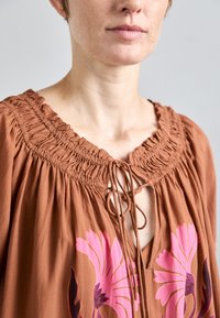 Braun Baumwollbluse mit einem gerafften Ausschnitt, Bindeschluss und großen rosa Blumenmustern auf der Vorderseite. Strukturierter, fließender Stoff.