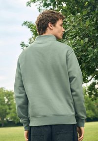 Sudadera verde con cuello alto, hecha de tejido suave; cuenta con puños y dobladillo acanalados, usada sobre pantalones oscuros en un entorno natural.
