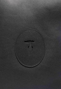 Svart lädersurface med en runt präglad logotyp och en central utskärningsdesign. Slät textur med subtila sömnadsdetaljer.