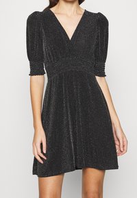 Svart klänning med V-urringning, korta puffärmar och figursydd midja. Tillverkad av ett skimrande material med en texturerad yta.