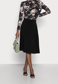 Falda negra plisada con cintura alta, combinada con una blusa floral de manga larga. Bolso verde con asa trenzada. Sandalias negras de punta abierta.