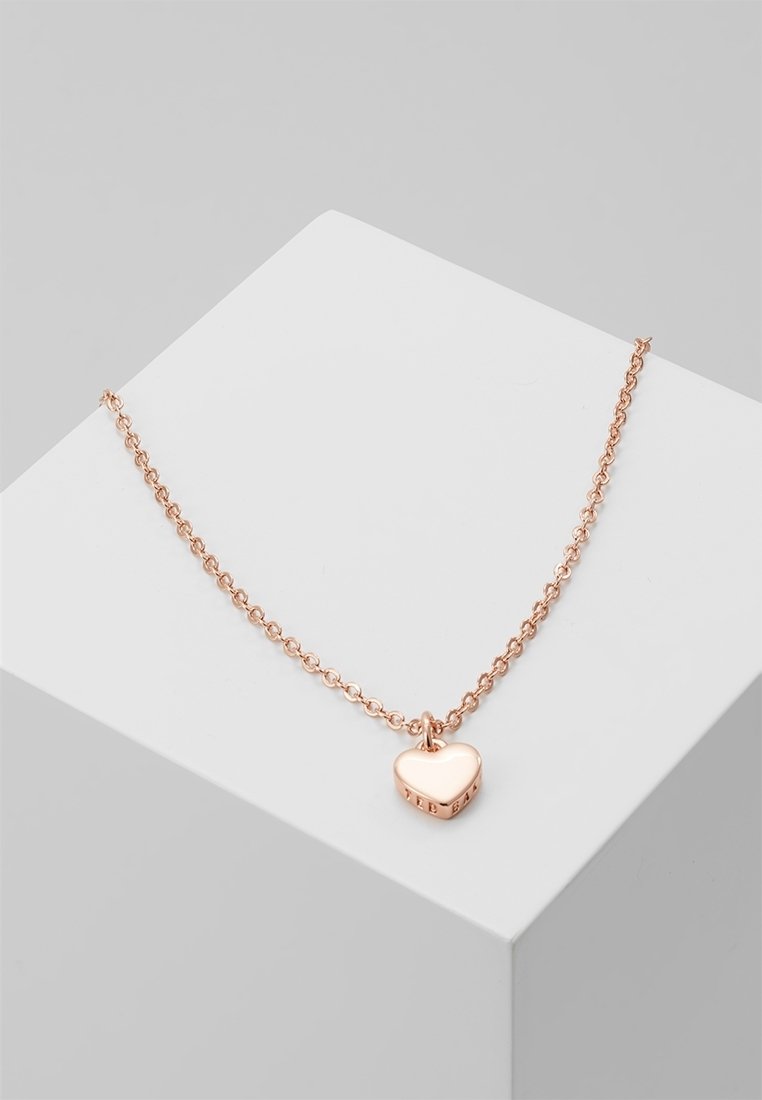 Collier chaîne en or rose avec un petit pendentif poli en forme de cœur reposant sur une surface géométrique blanche.