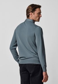 Herrenpullover aus weichem, atmungsaktivem Stoff. Hellblau-graue Farbe, gerippte Bündchen und Saum, klassischer Kragen, figurbetonte Passform. Schwarze Hose sichtbar darunter.