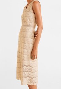 Robe beige sans manches avec motif en dentelle ondulée, taille cintrée et ourlet à hauteur du genou. Texture lisse avec des détails découpés.