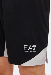 Μαύρη και γκρι βερμούδα με το λογότυπο EA7 Emporio Armani στο κάτω αριστερό πόδι που φοριέται από ένα άτομο.
