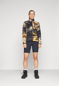 Fleece jaka melnā, bēšā un oranžā tūku rakstā, ar rāvējslēdzēju priekšā, apvienota ar tumšām šortiem un melniem pārgājienu apaviem.