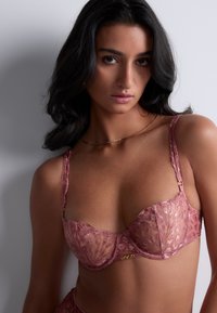 Soutien-gorge en dentelle rose doux avec un motif floral transparent, support à armatures, bretelles réglables et accents en métal doré.