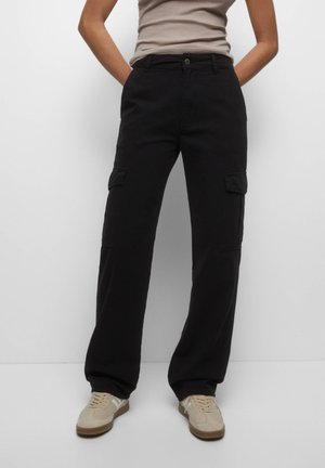 Personne portant un pantalon cargo noir avec des poches et des baskets beiges, se tenant les mains dans les poches contre un fond uni.