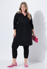 Robe noire à manches longues avec un décolleté en V et une taille froncée, associée à des leggings noirs et des ballerines en suédine rose. Tenant une pochette rose.
