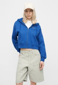 Νεαρό άτομο με ίσια ξανθά μαλλιά, που φοράει λευκό καπέλο, μπλε φούτερ με φερμουάρ, λευκό πουκάμισο και ανοιχτό γκρι σορτς μέχρι το γόνατο, στέκεται μπροστά σε απλό φόντο.