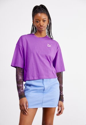 Camiseta de algodón morada de manga corta con cuello redondo. Presenta un pequeño logo blanco de Puma. Combinada con una minifalda de mezclilla azul clara.